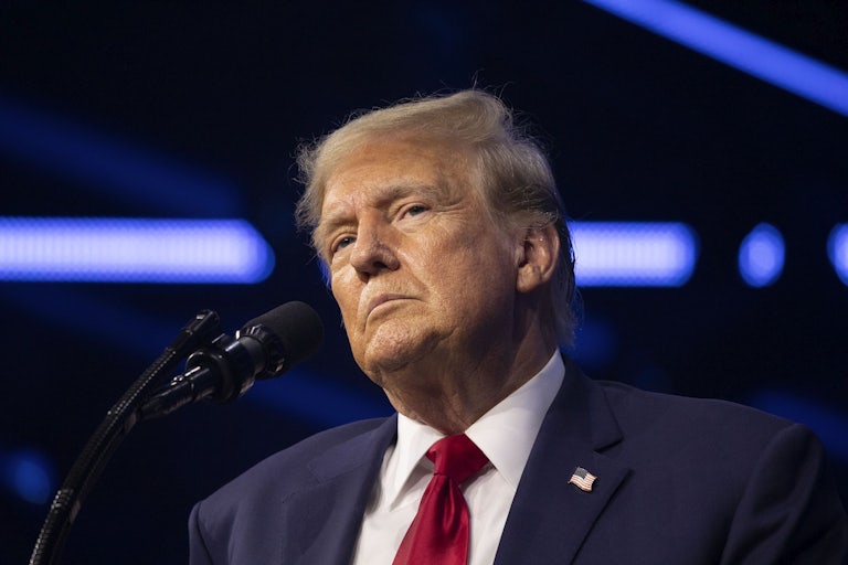 Donald Trump stands before a mic, head tilted. He looks serious.