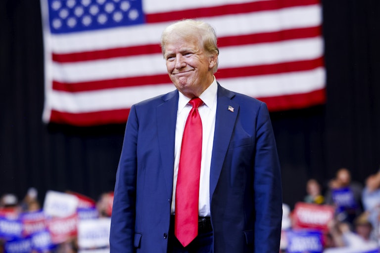Donald Trump toothlessly smiles as he stands before a large American flag at a political rally